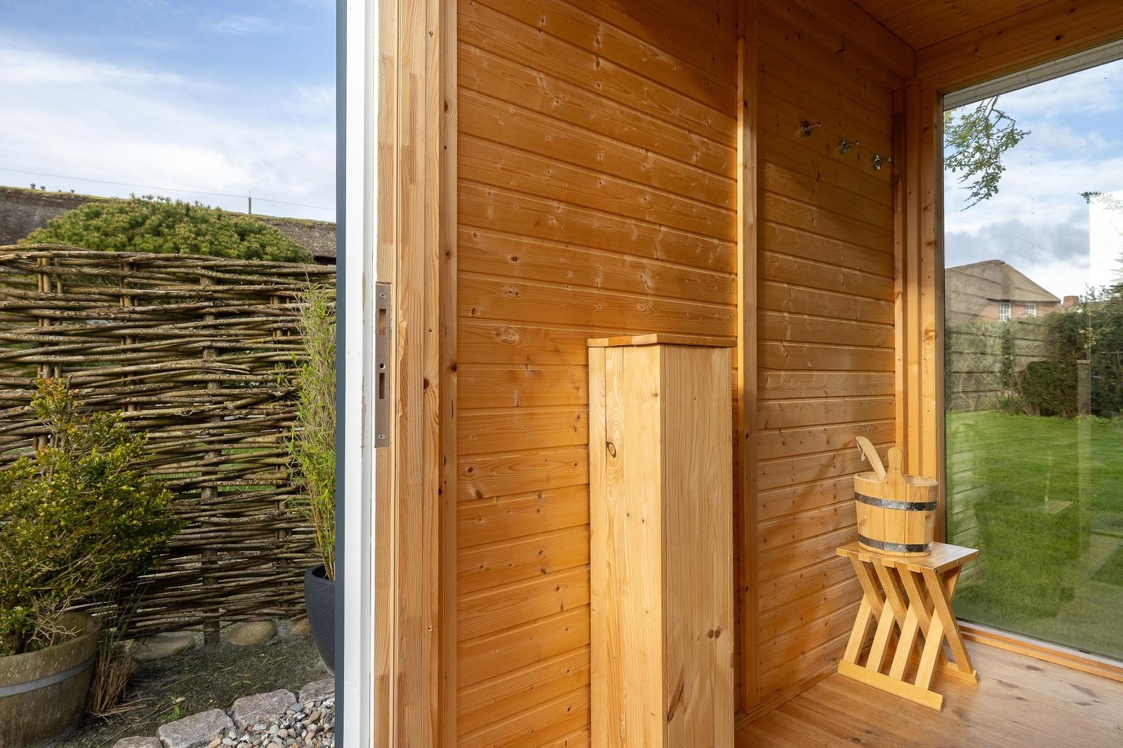 Holzsauna mit Einstiegsbereich und Außentür. Holzbank und Eimer stehen bereit.