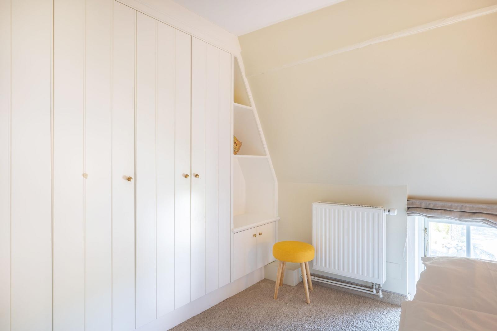 Schlafzimmer mit weißem Schrank, gelbem Hocker und Fenster.