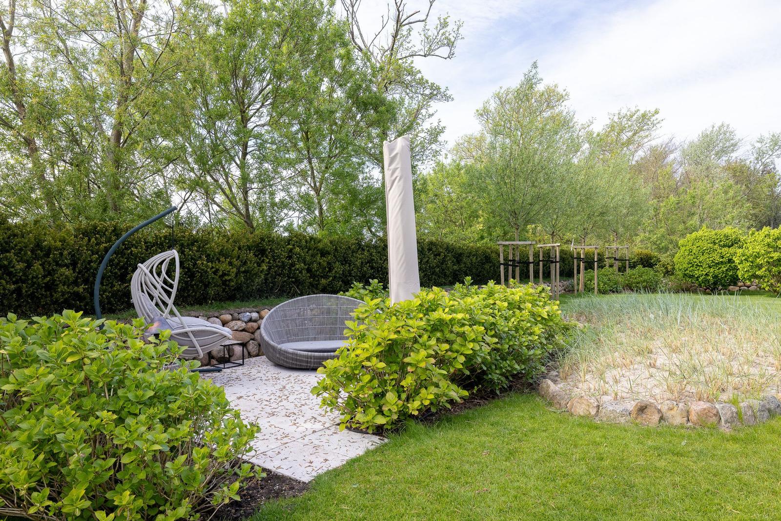 Garten mit Sitzbereich, Grasfläche und Sträuchern. Hintergrund: Bäume und Hecke.