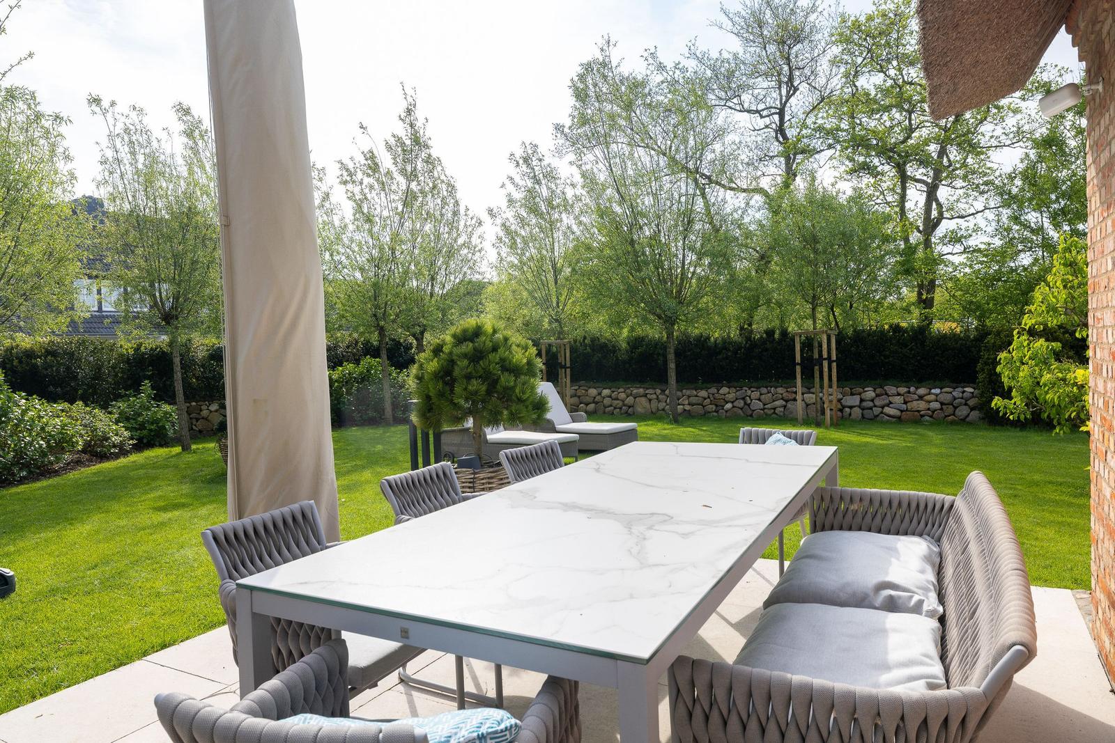 Terrasse mit Tisch und Sitzgruppe, umgeben von Grünfläche und Bäumen.