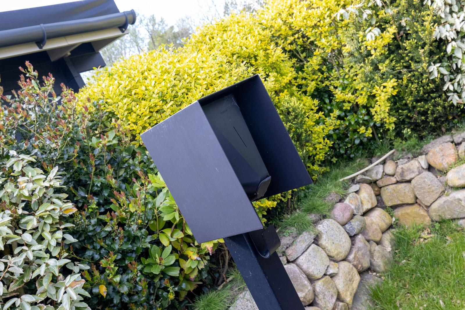 Schwarzer Briefkasten im Garten neben Sträuchern und Steinmauer.