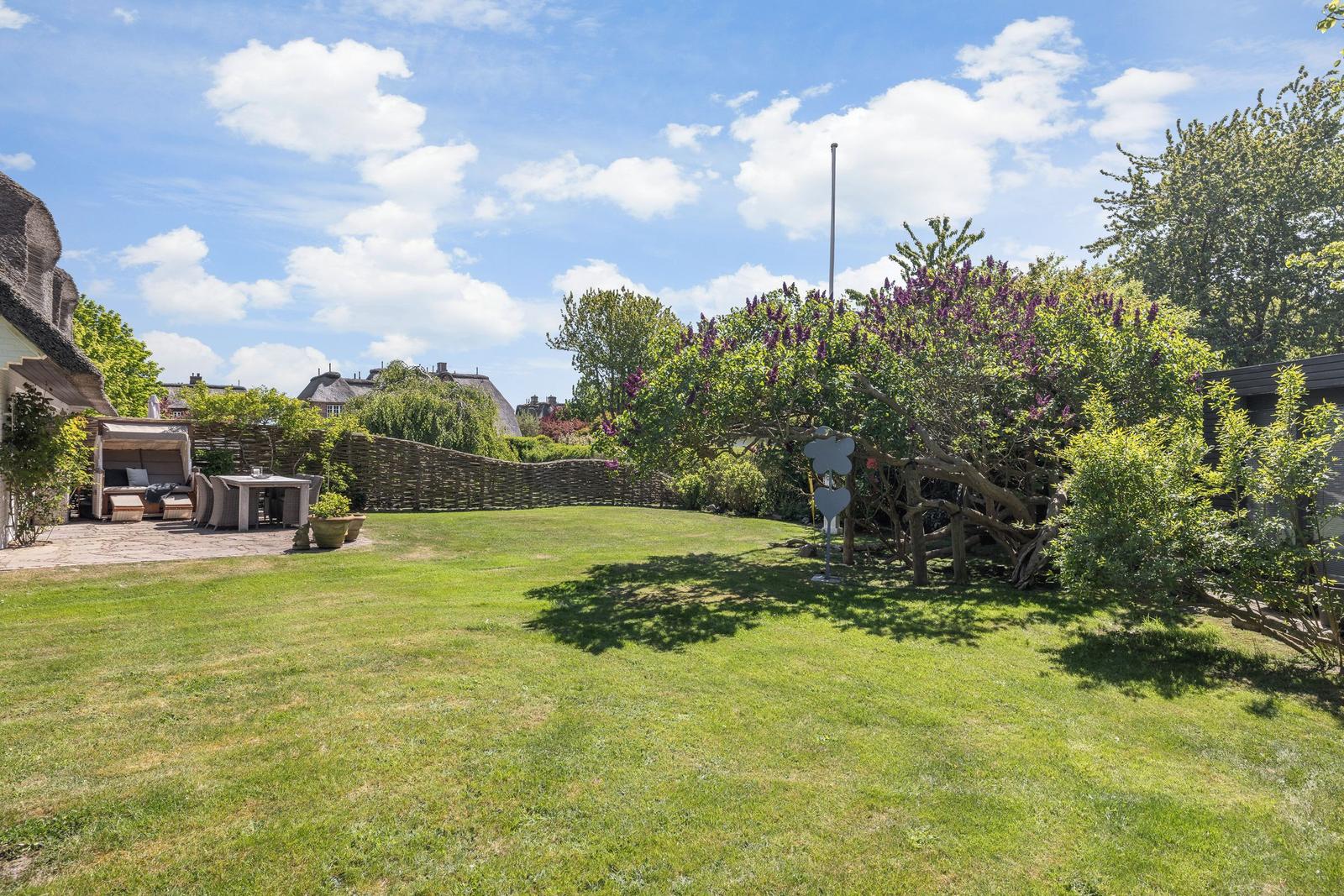 Green garden with terrace, flowers, and trees under blue sky.