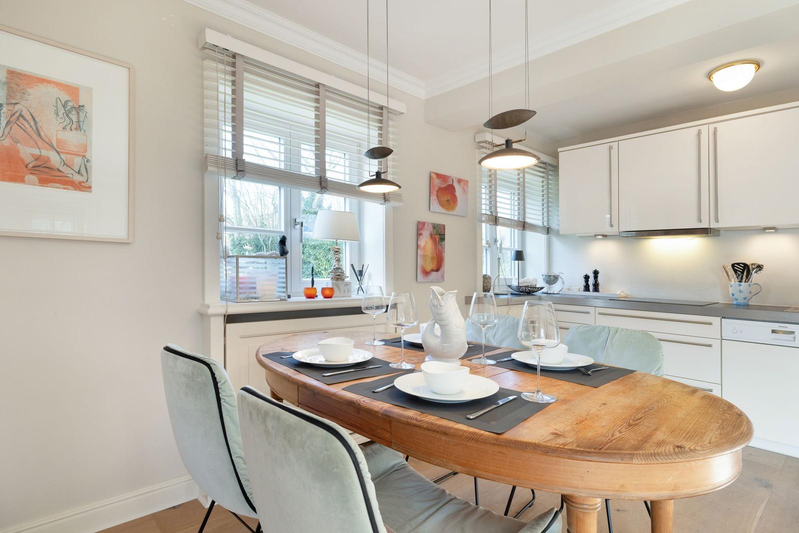 Dining room with wooden table, chairs, and windows. Kitchen with white cabinets.