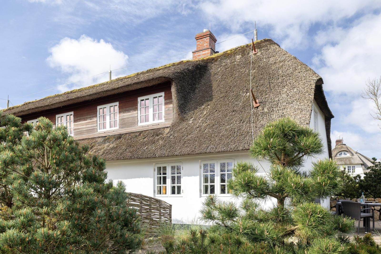 Haus mit strohgedecktem Dach, weißen Wänden und Fenstern, umgeben von Bäumen.
