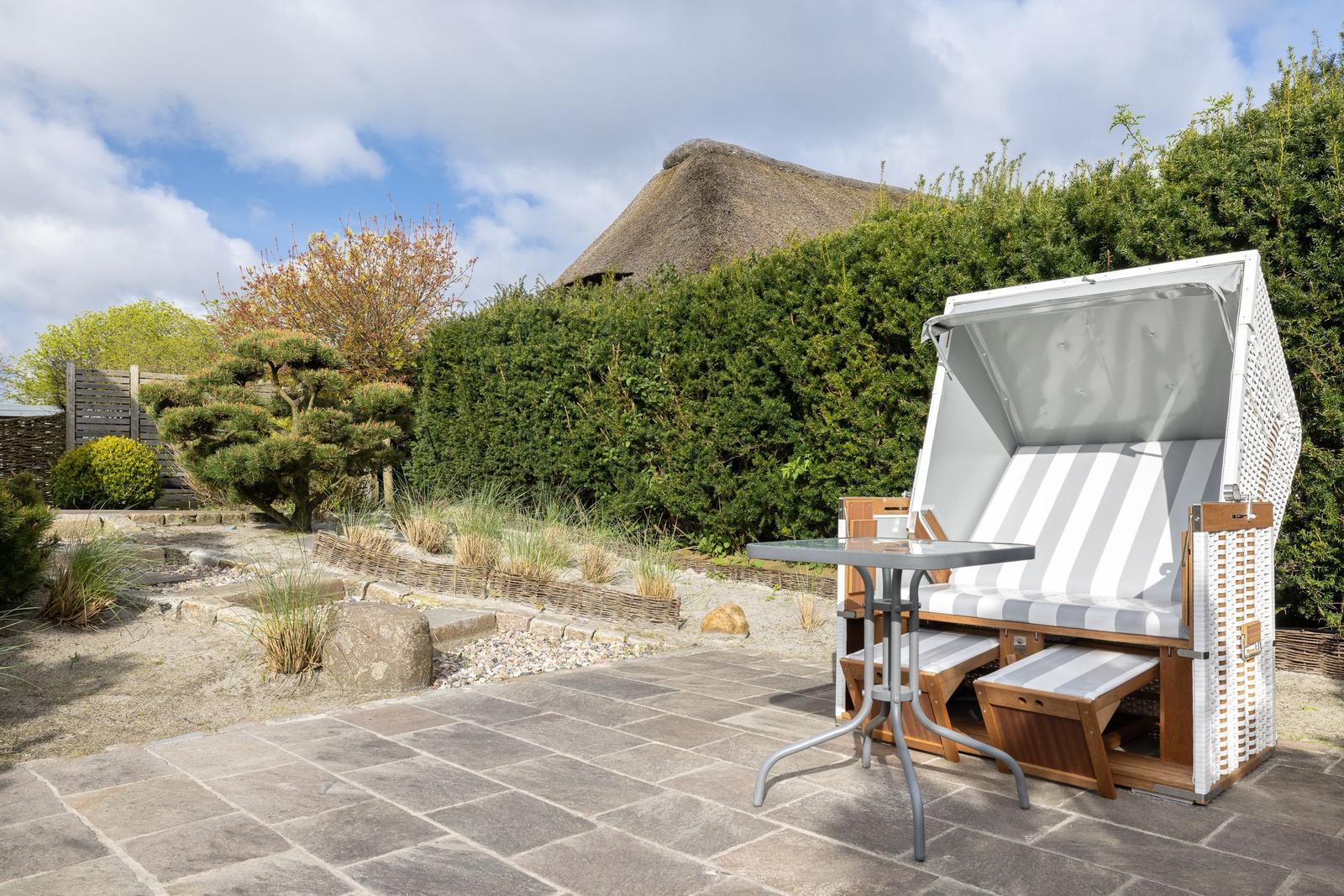 Terrasse mit Strandkorb und Tisch, Hintergrund mit Hecke und Strohdach.