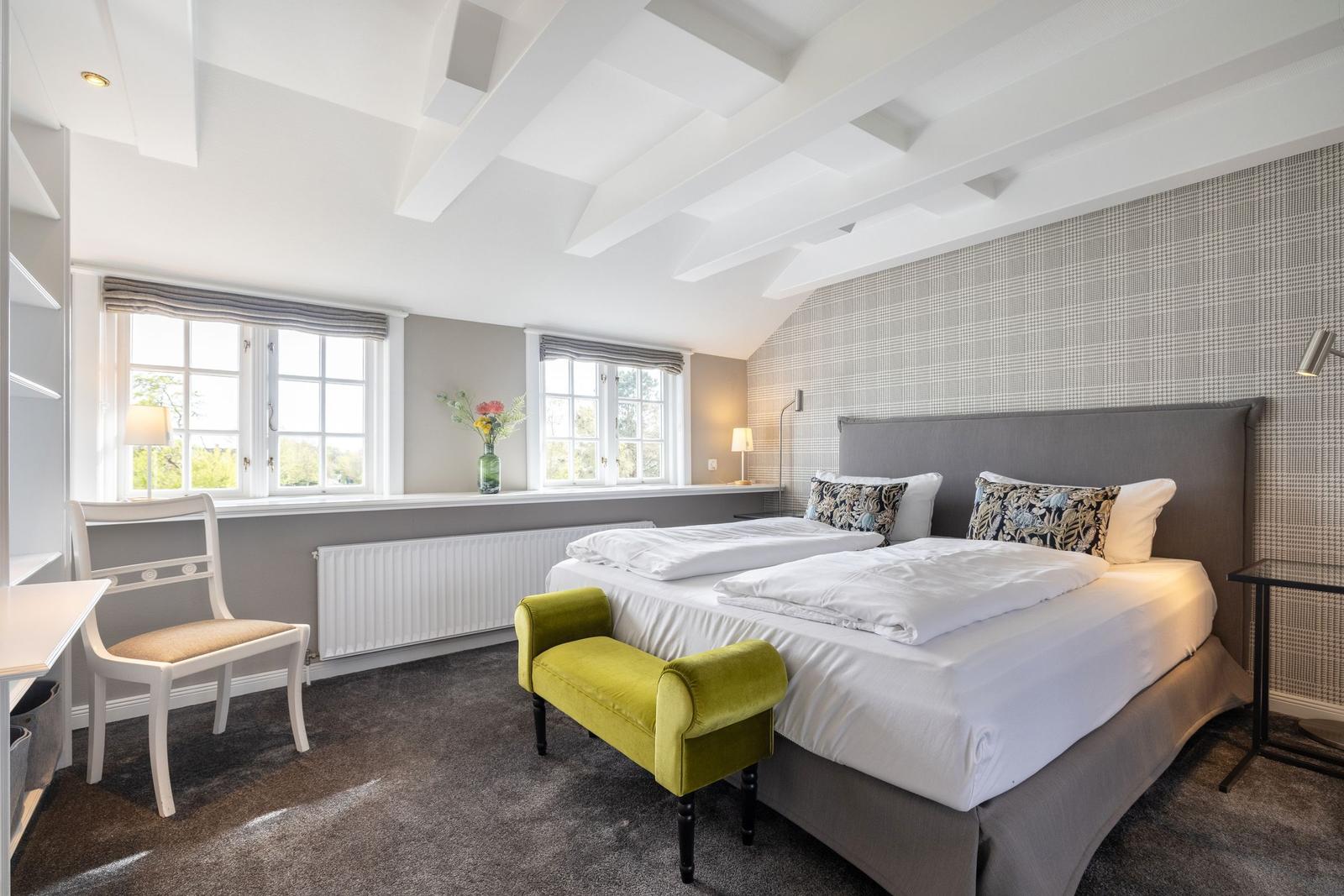 Schlafzimmer mit Doppelbett, grünem Hocker und Fenstern mit Blick nach draußen.