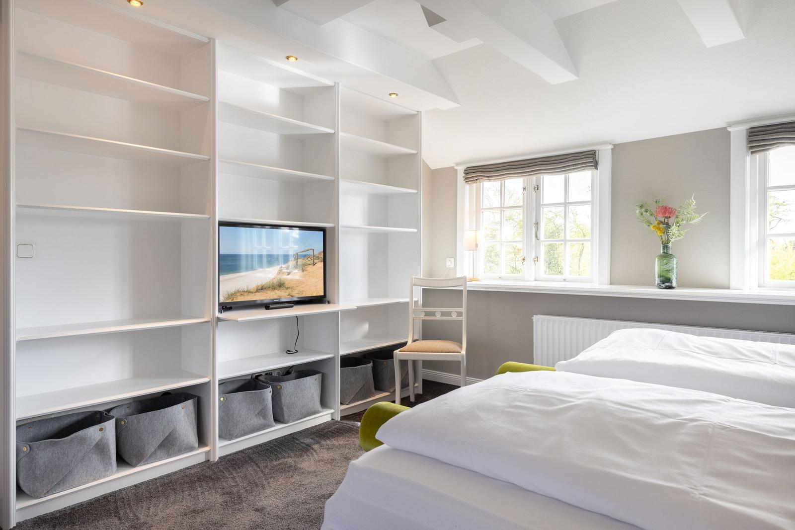 Schlafzimmer mit Bett, TV und Fenster. Weiße Regale und grauer Teppich.