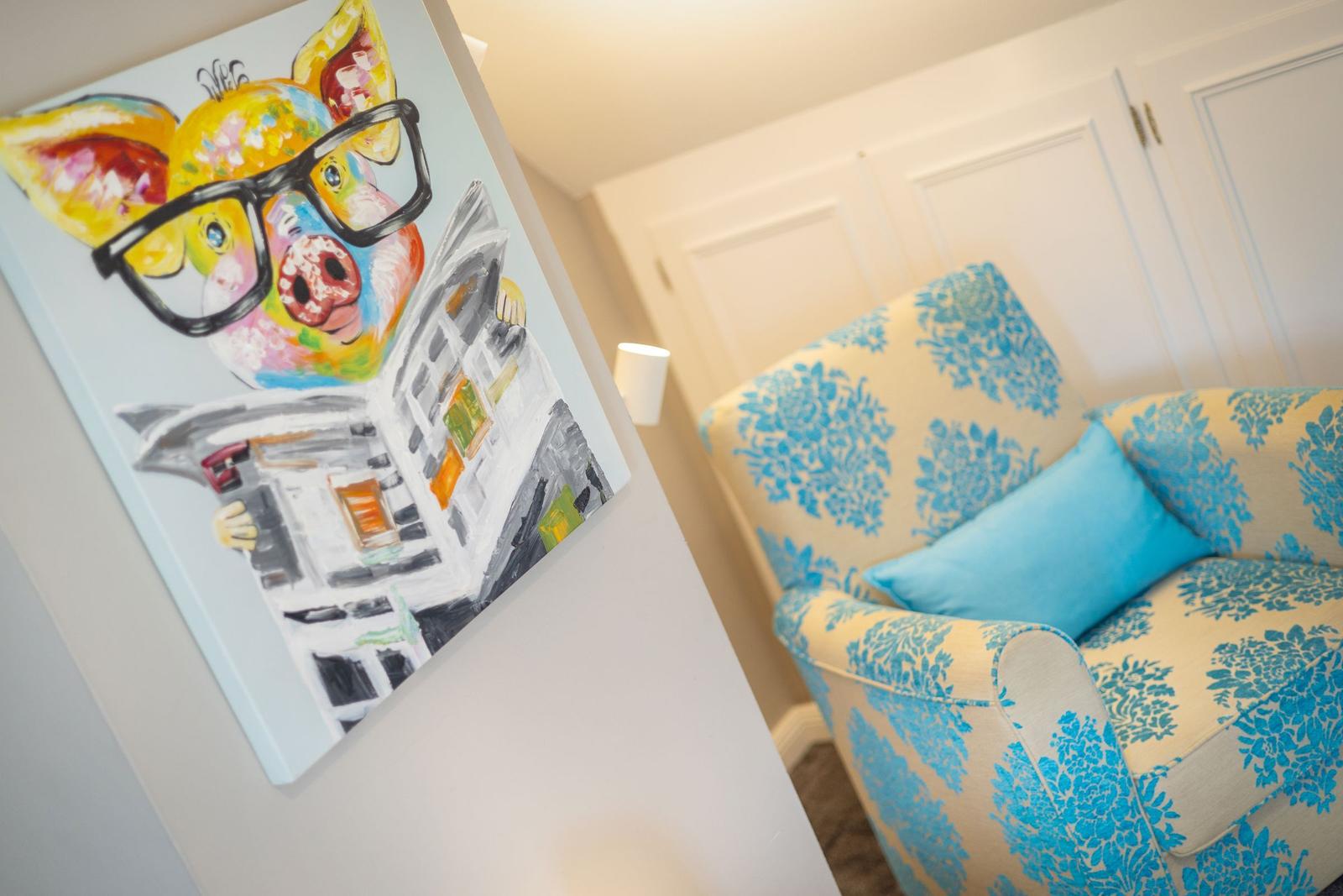 Wandbild mit Schwein in Brille neben einem Sessel mit Blumenmuster.