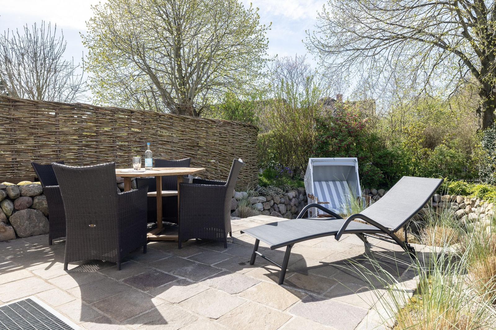 Terrasse mit Tisch, Stühlen und Liegen. Hintergrund: Holzzaun und Grünflächen.