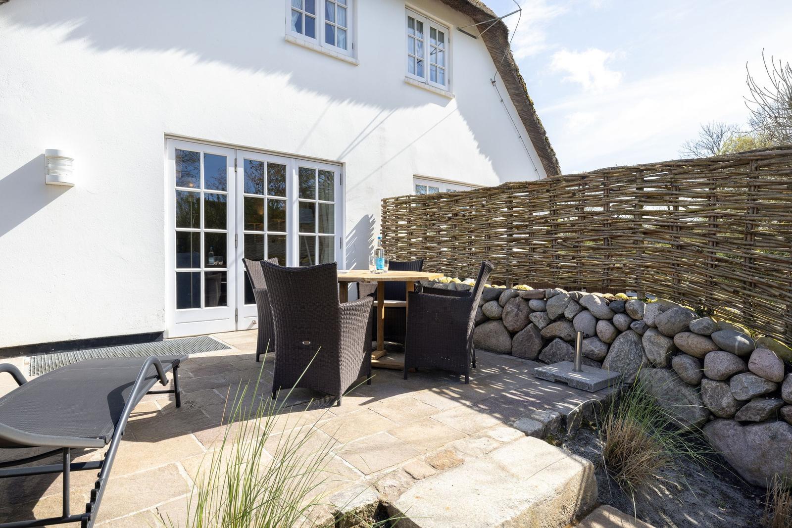 Terrasse mit Tisch, Stühlen und Liegestuhl vor weißem Haus mit Schiebetür.