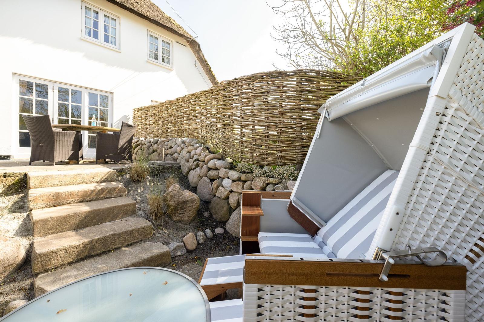 Weißer Strandkorb mit Streifenkissen im Garten vor einem Haus mit Steintreppe.