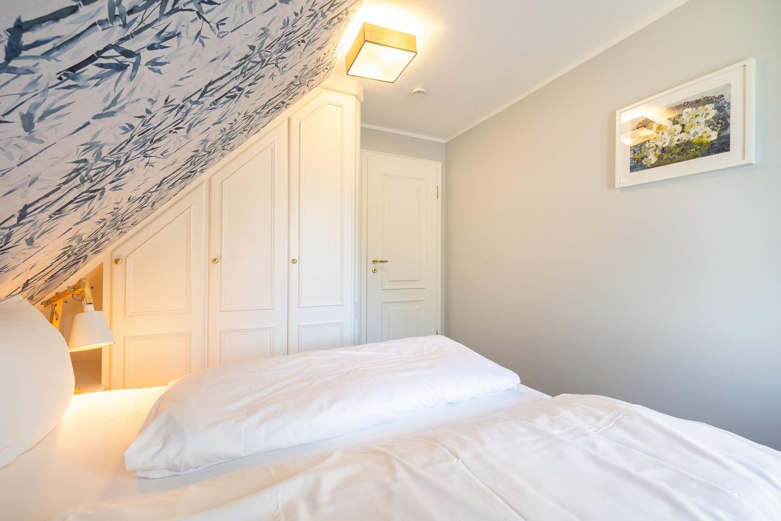 Schlafzimmer mit weißem Bett, Kleiderschrank und gemusterter Decke.