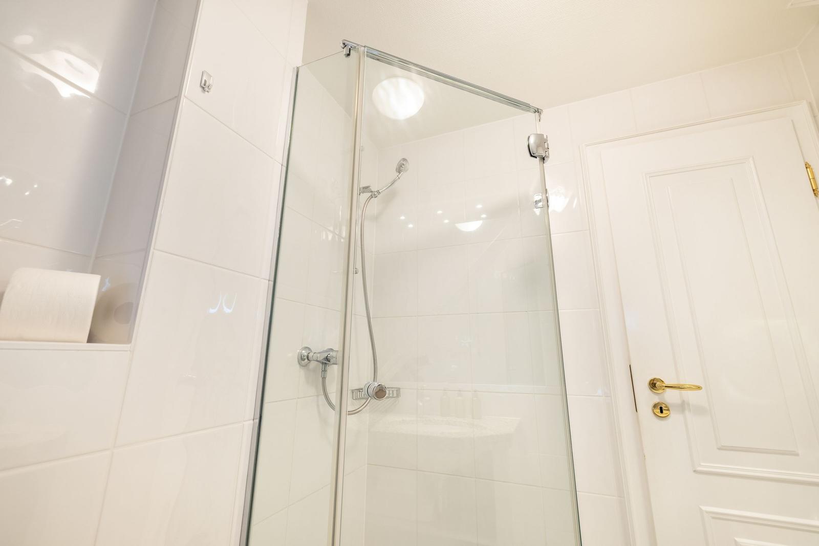 Dusche mit Glaswand, weiße Fliesen und goldener Türknauf.