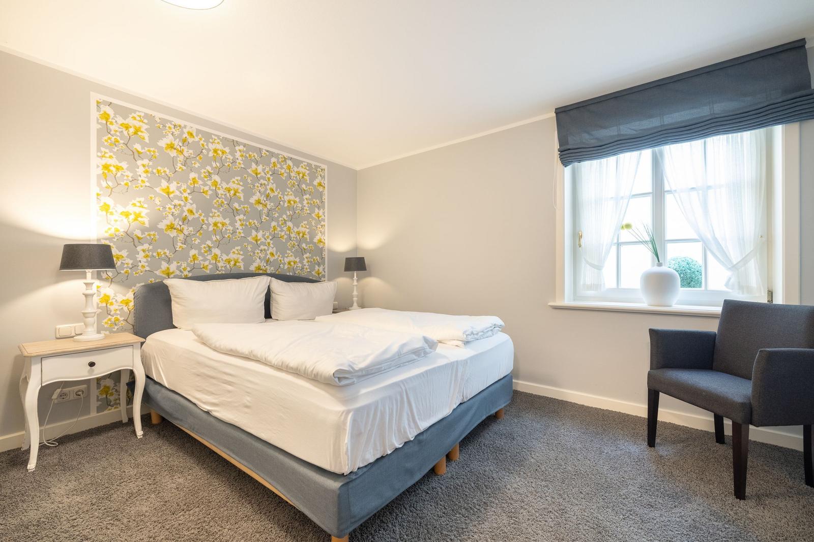 Schlafzimmer mit Bett, Nachttisch und Stuhl. Wand mit Blumenmotiv. Fenster mit Vorhängen.