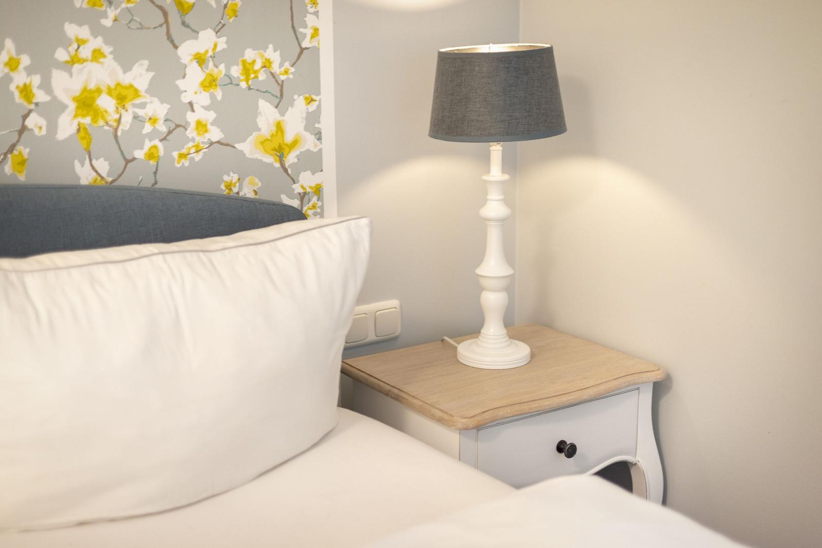 Bettkopfteil mit Kissen, Nachttisch mit Lampe und floraler Tapete.