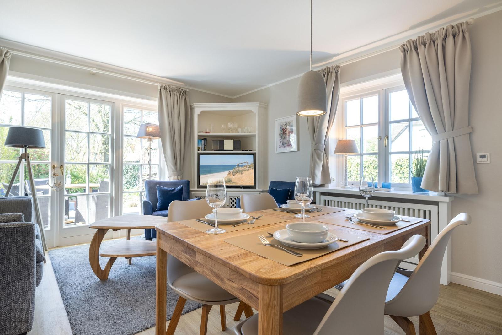 Esszimmer mit Holztisch, Stühlen und Fenstern. Fernseher und Wohnbereich sichtbar.