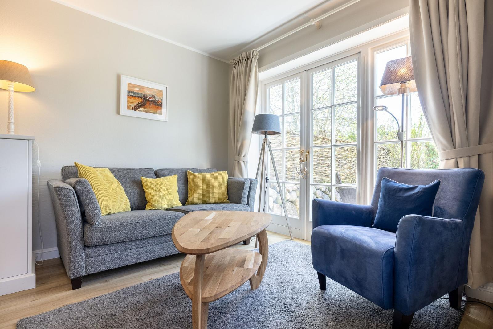 Wohnzimmer mit grauem Sofa, blauem Sessel und Holztisch. Fenster mit Blick auf Außenbereich.