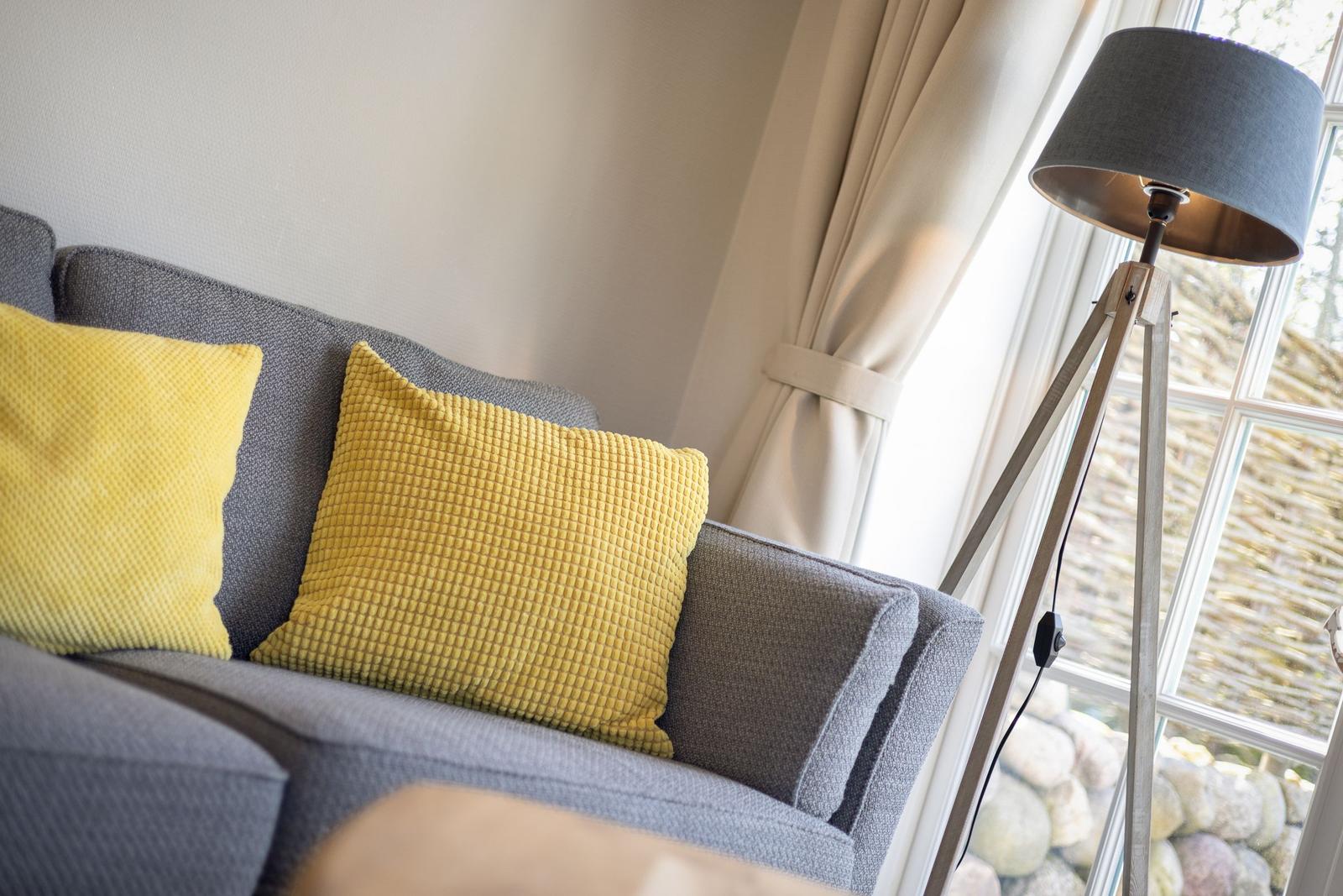 Ein grauer Sofa mit zwei gelben Kissen und einer Stehlampe neben einem Fenster.