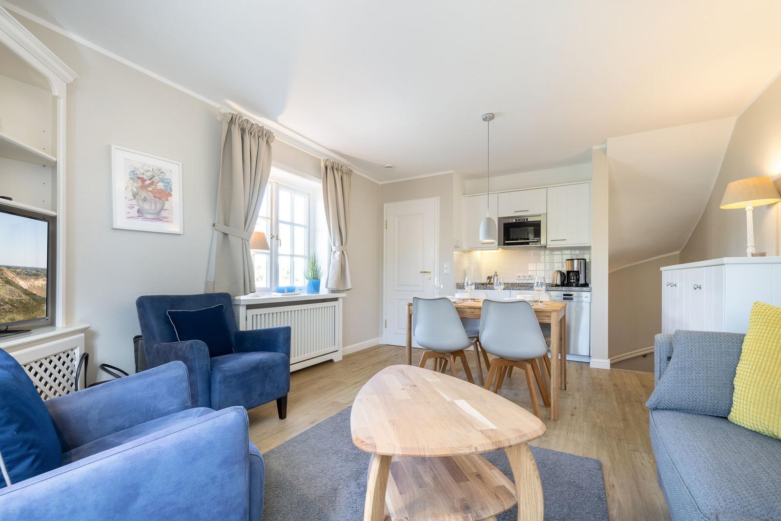 Wohnzimmer mit Essbereich und Küche. Blaue Sessel, Holztisch, Fenster mit Vorhängen.