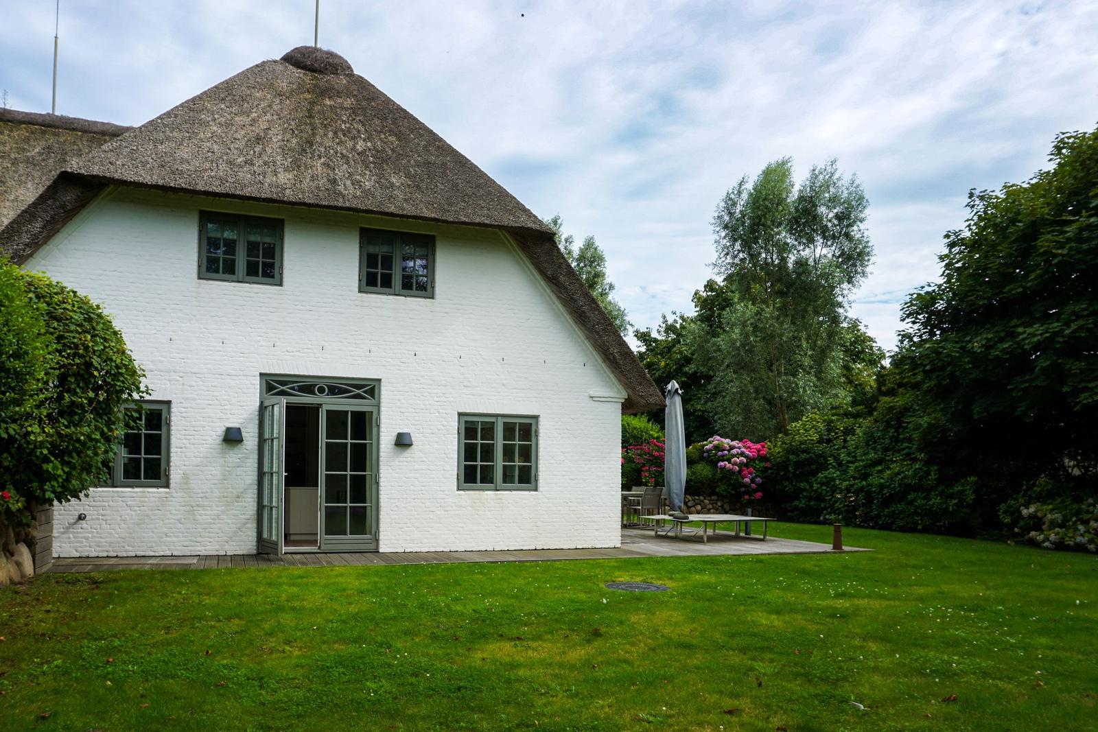 White house with thatched roof, garden and terrace with table and chairs.