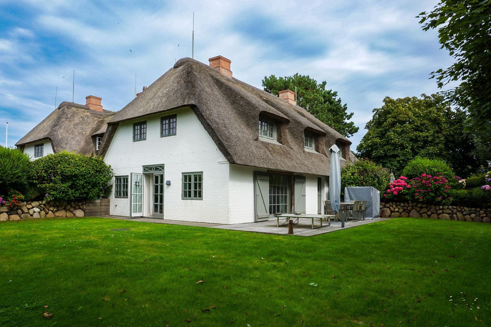 House with thatched roof, terrace, and garden. Wooden tables and chairs outdoors.