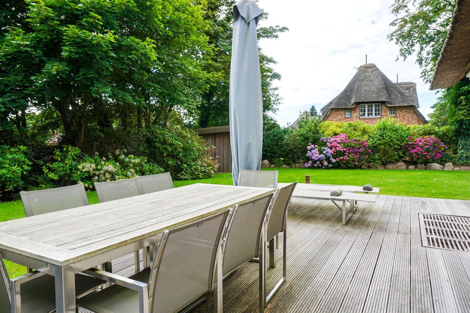Wooden terrace with table, chairs, and umbrella. A house with thatched roof in the background.