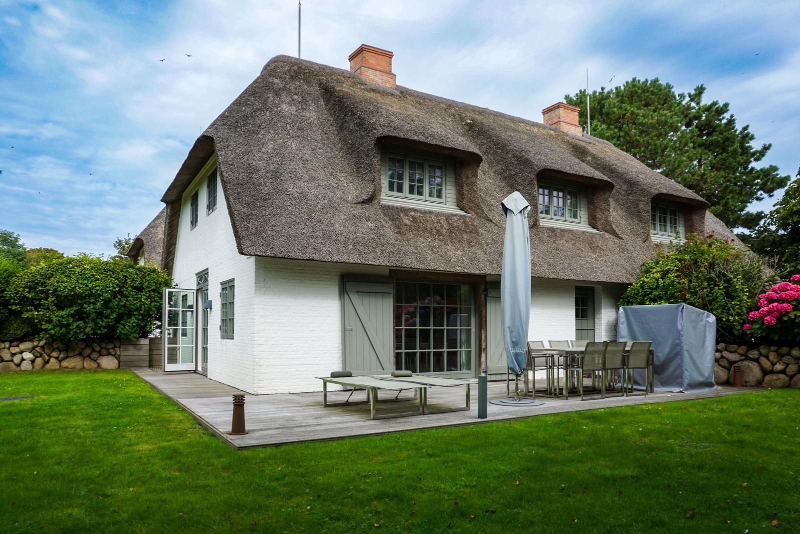 House with thatched roof, terrace, and garden. Table, chairs, and umbrella are outside.