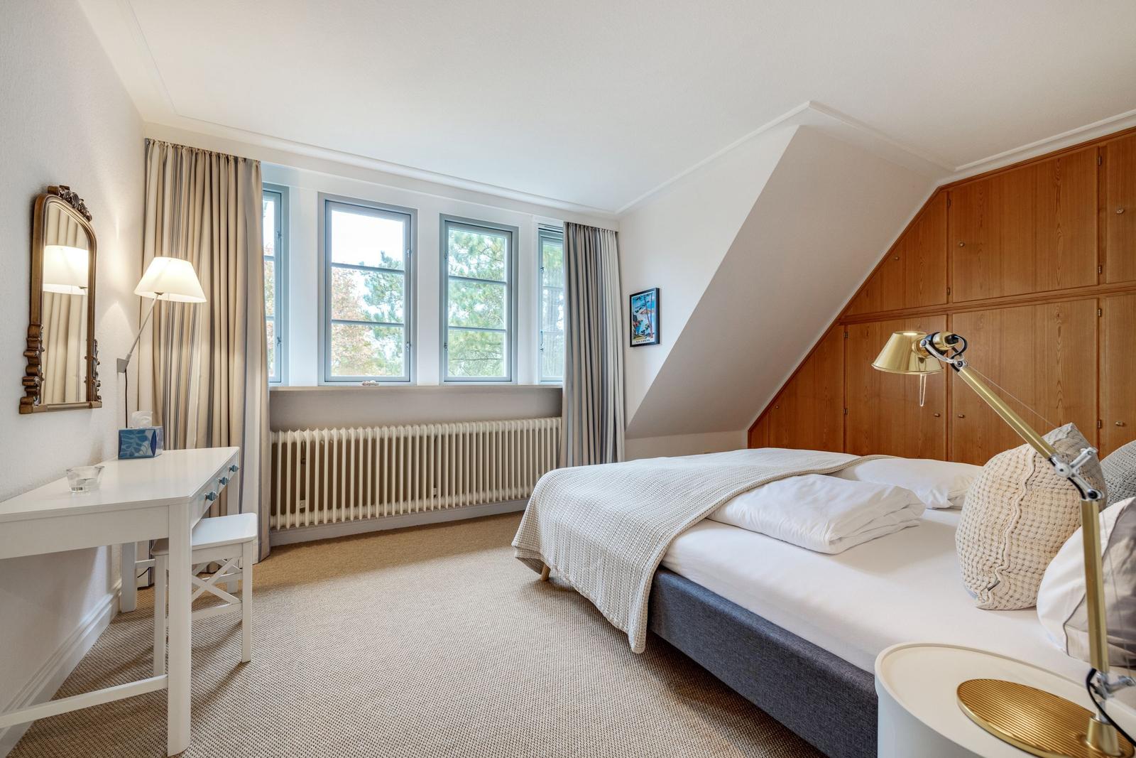 Schlafzimmer mit Bett, Schreibtisch und Fenstern. Holzschrank an der Wand.