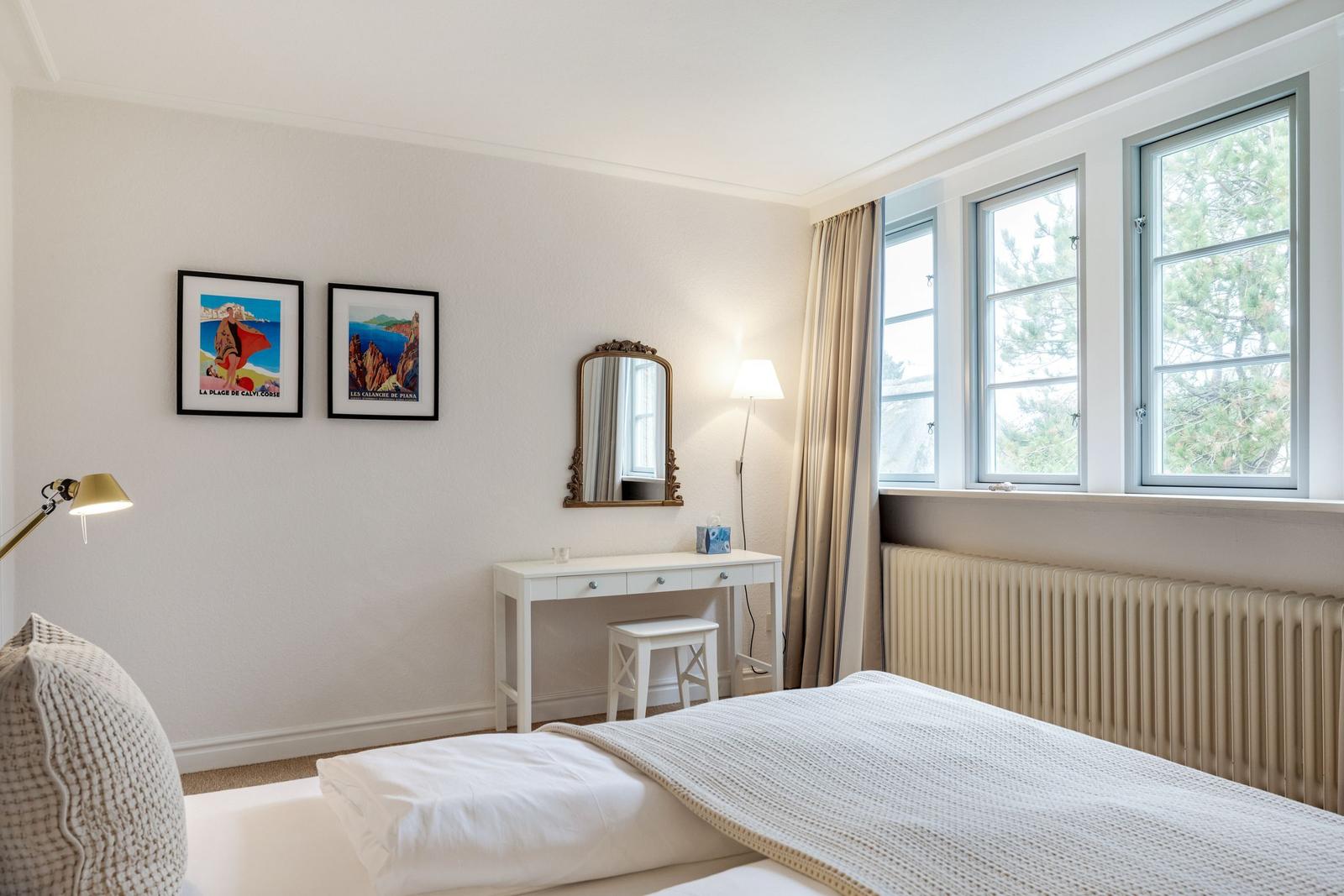 Schlafzimmer mit Bett, Schreibtisch und Fenstern mit Blick auf Bäume.