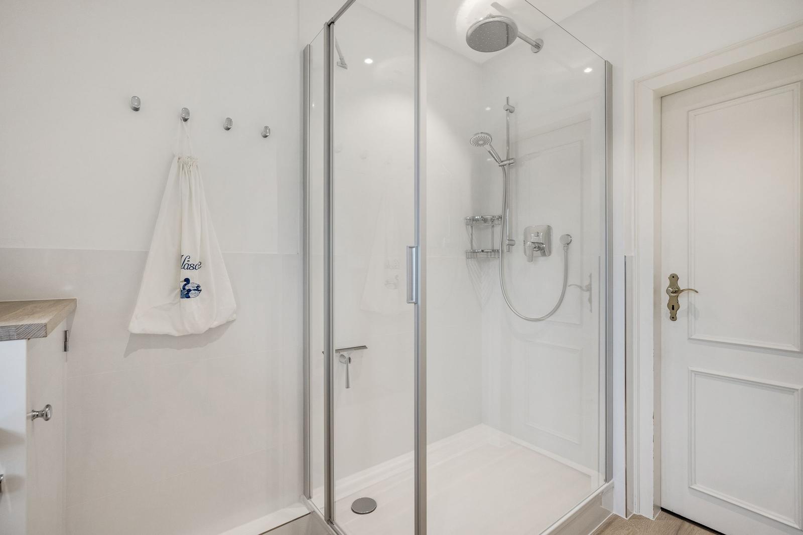 Dusche mit Glaswand, Duschbrause und Handbrause. Wand mit Haken und weißer Tasche.