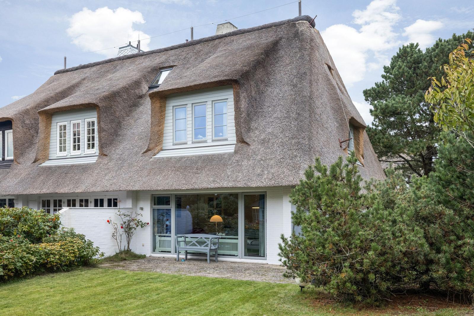 Haus mit strohgedecktem Dach und weißen Wänden, umgeben von Grünflächen und Bäumen.