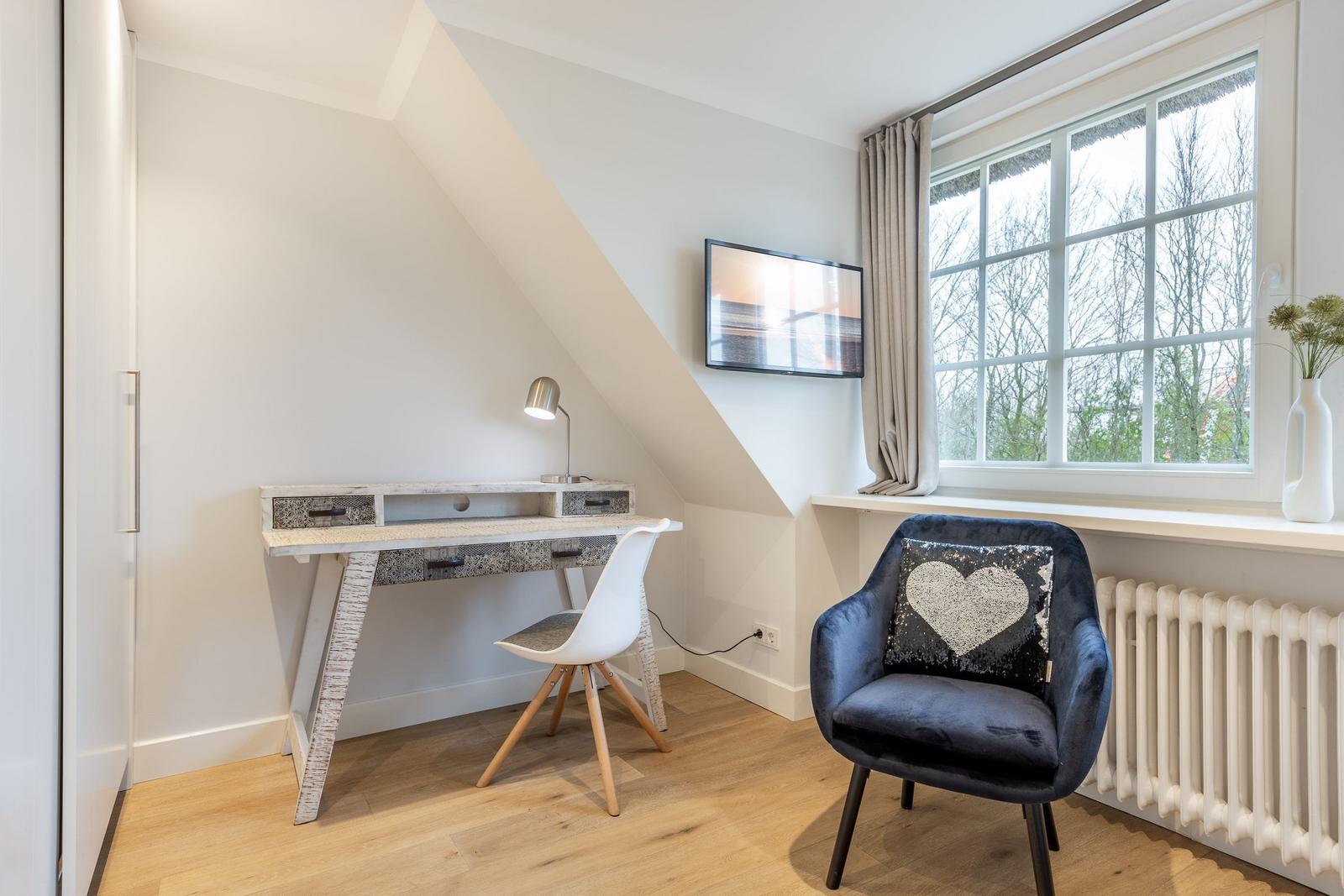 A bedroom with a desk, chair, and armchair by a window.