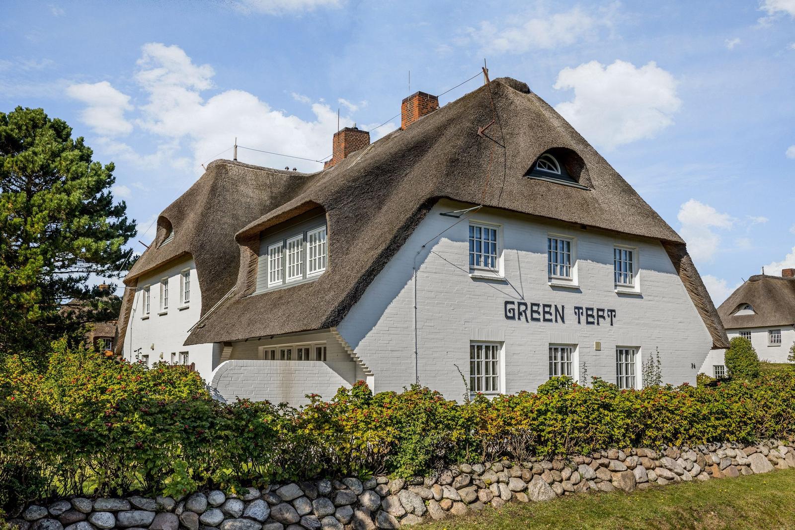 Weiße Hütte mit Strohdach und Gartenzaun. Schriftzug GREEN TEFT auf Fassade.