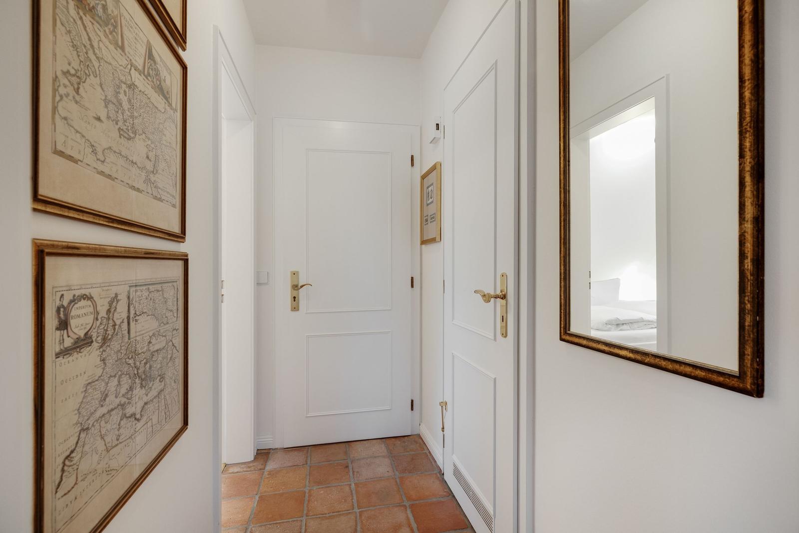 Flur mit weißen Türen, Terrakottaboden und gerahmten Karten an der Wand.