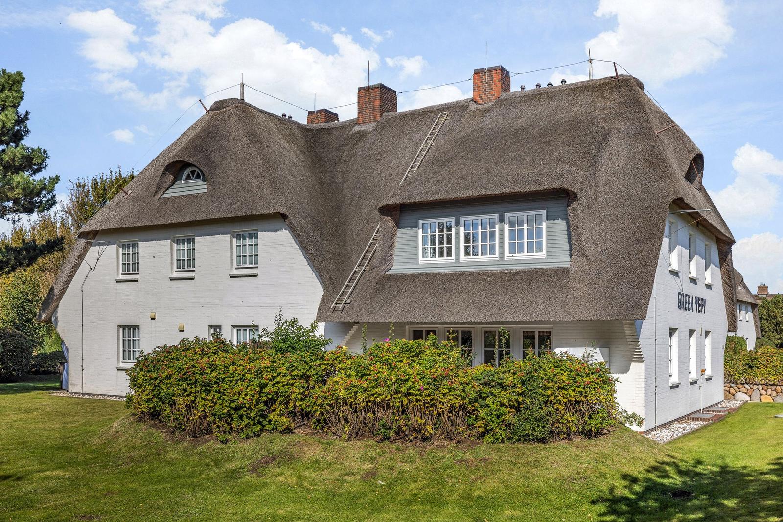 Traditionelles Haus mit Strohdach und weißen Wänden, umgeben von Grünflächen und Sträuchern.
