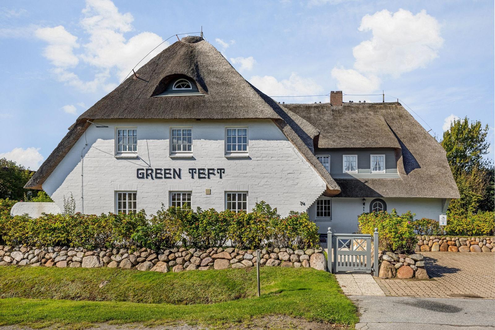 Weißes Haus mit Strohdach und Gartenzaun. Schriftzug GREEN TEFT auf Fassade.