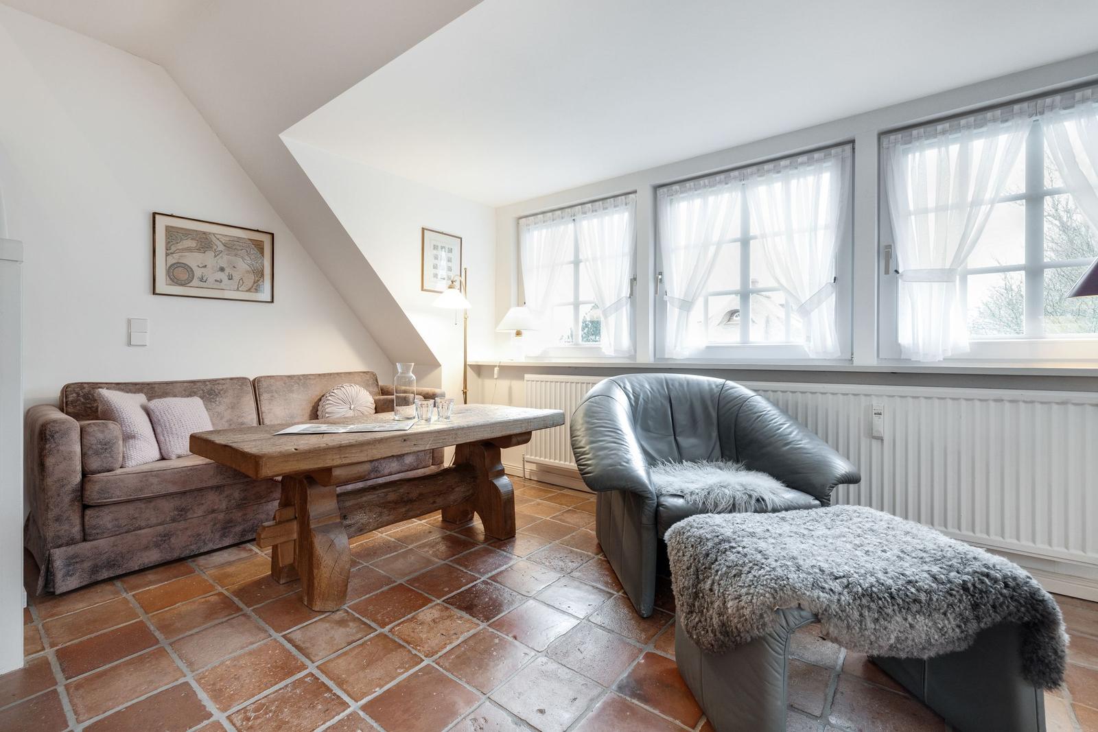 Wohnzimmer mit Sofa, Holztisch und Ledersessel. Terrakottaboden und Fenster mit Vorhängen.