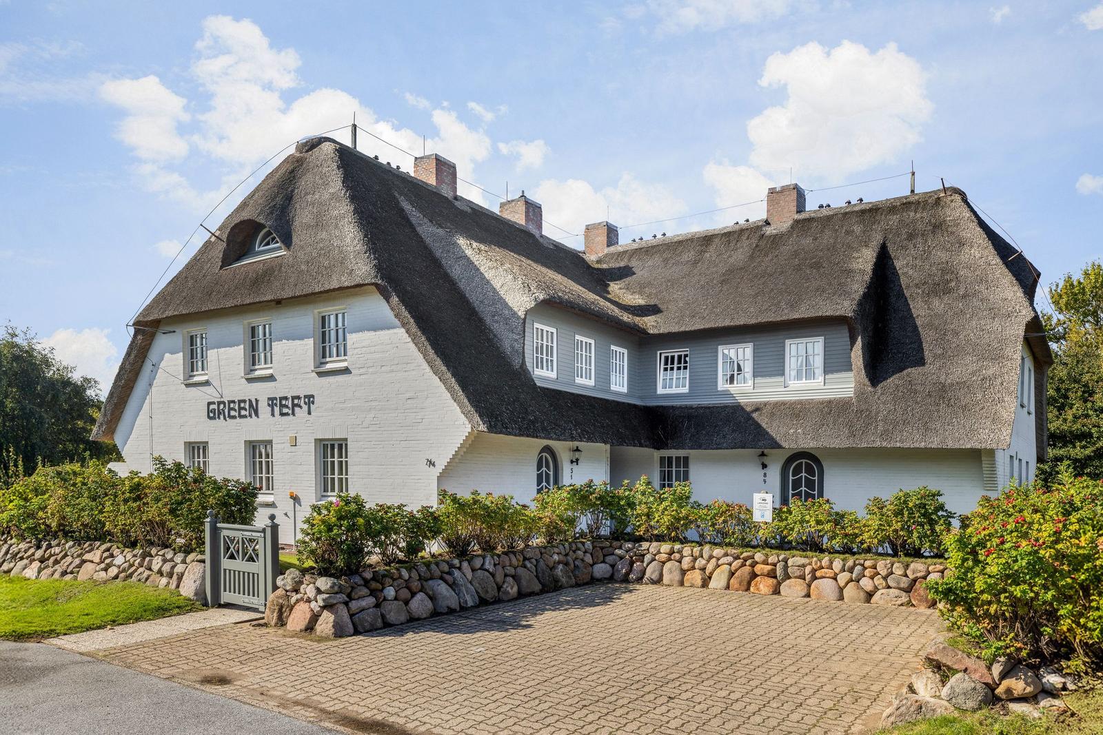 Ein großes Haus mit Strohdach und weißen Wänden. Vorgarten mit Steinmauer und Pflanzen.