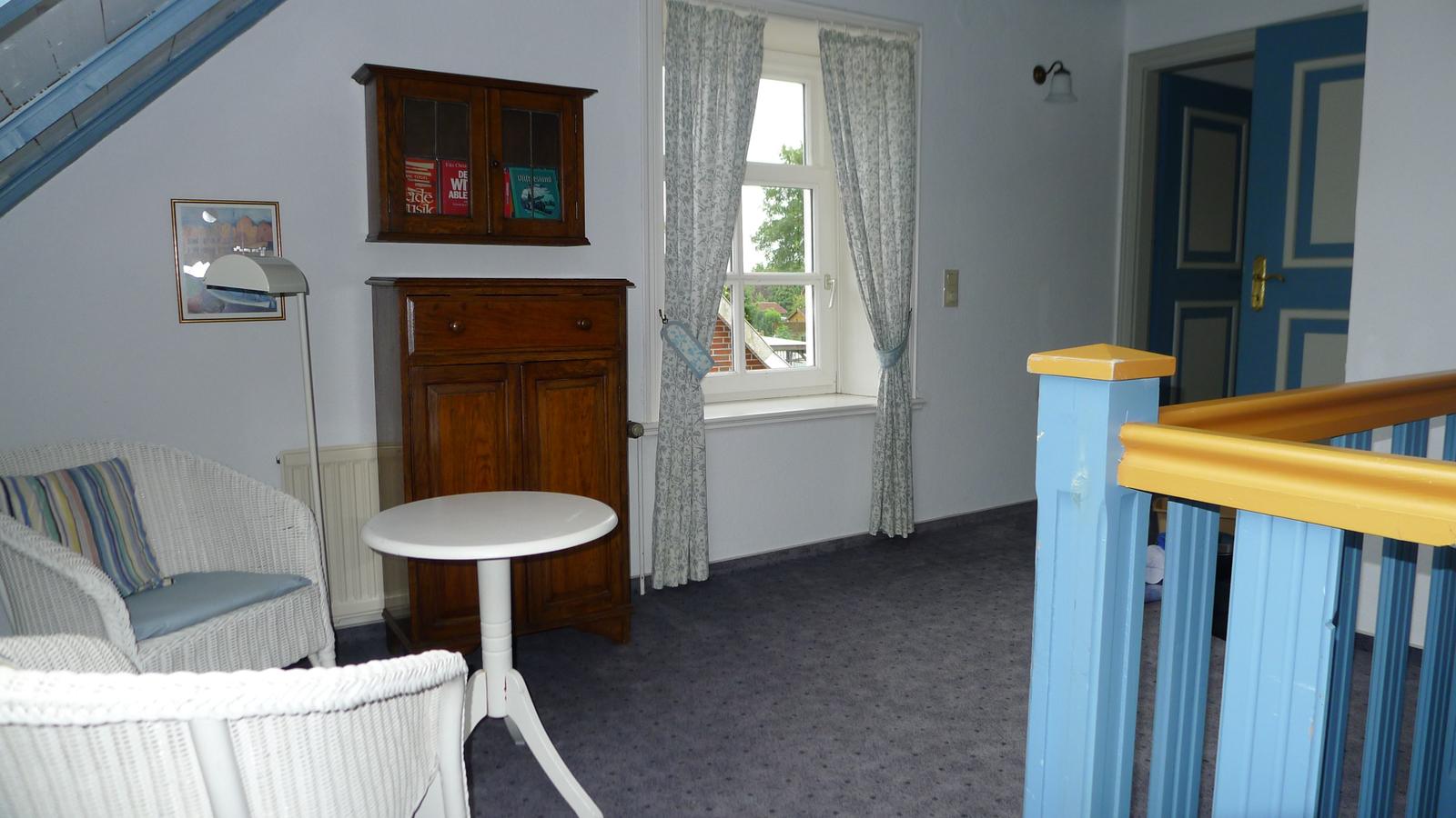 Zimmer mit Holzschrank, Fenster, Gardinen und Wicker-Sofa. Gelb-blauer Geländer im Vordergrund.