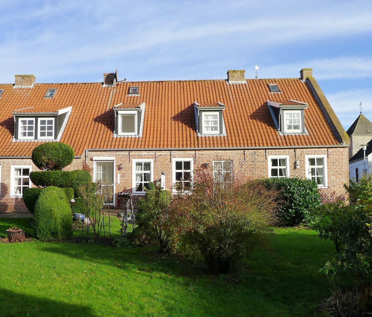 Haus mit rotem Ziegeldach und Garten. Weiße Fenster und Schornsteine sind sichtbar.