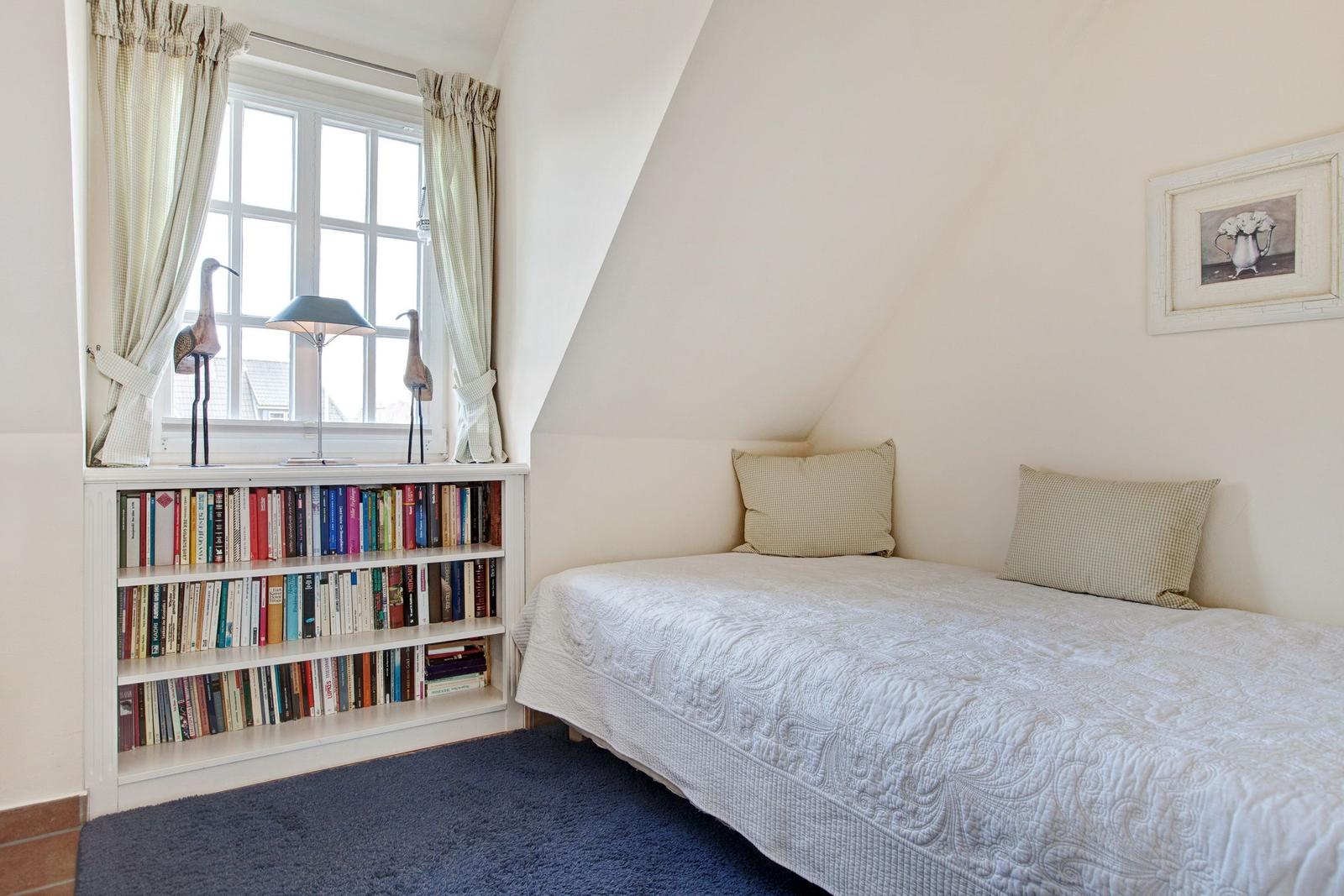 A bedroom with sloped ceiling, bed, bookshelf, and window with curtains.