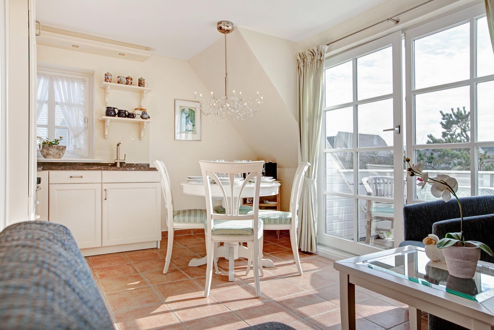 Dining room with white table, chairs, and kitchen area. Large windows with view to balcony.