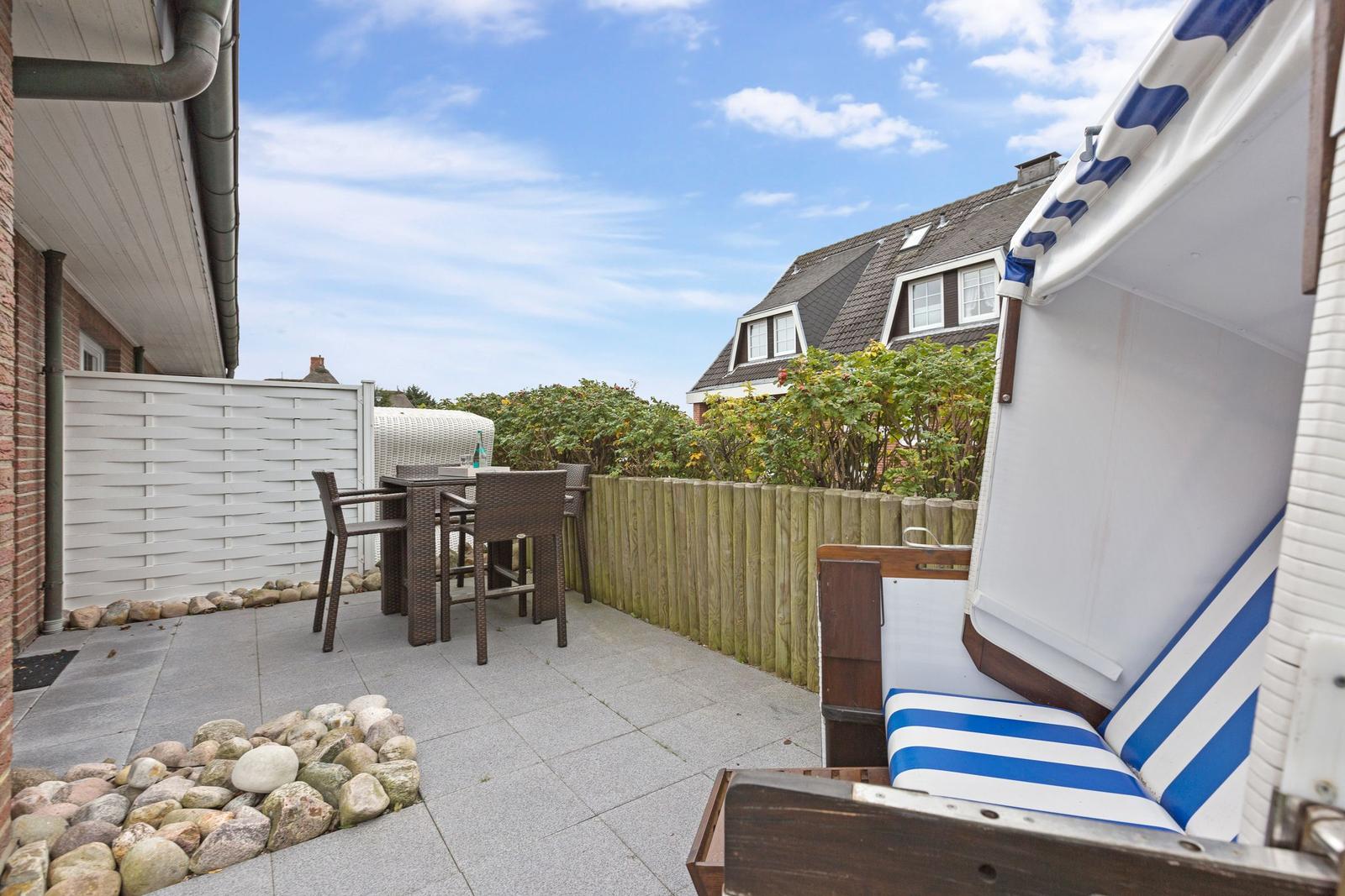 Terrasse mit Strandkorb, Tisch und Stühlen. Hinterhof mit Holzzaun und Pflanzen.
