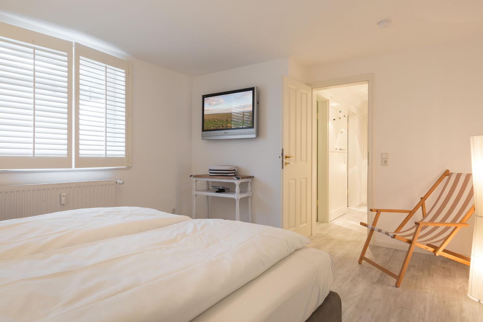 Zimmer mit Doppelbett, TV und Liegestuhl. Fenster mit Jalousien. Tür zum Bad.