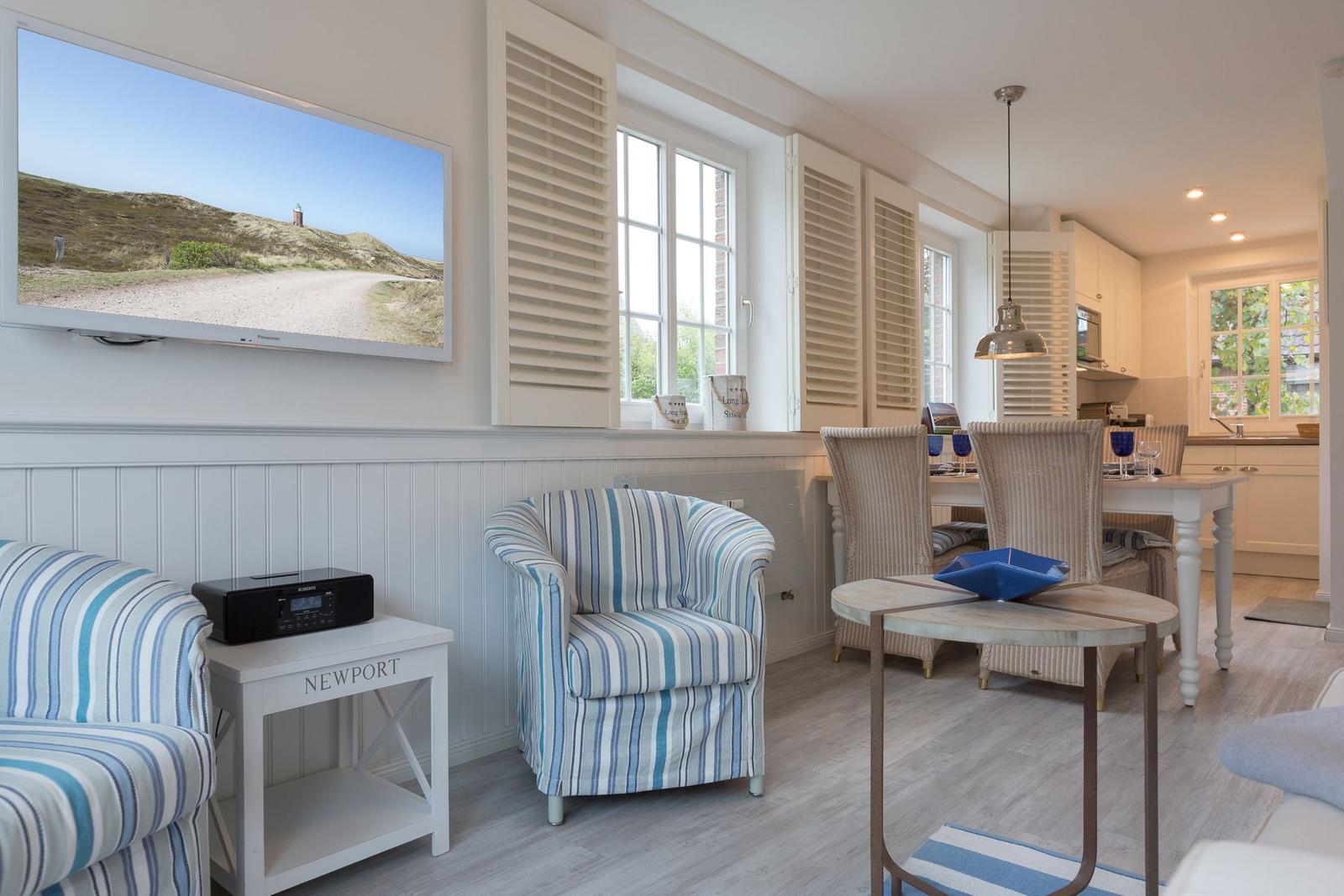 Wohn- und Essbereich mit TV, Stühlen und Tisch. Fenster mit Jalousien und Küchenbereich im Hintergrund.