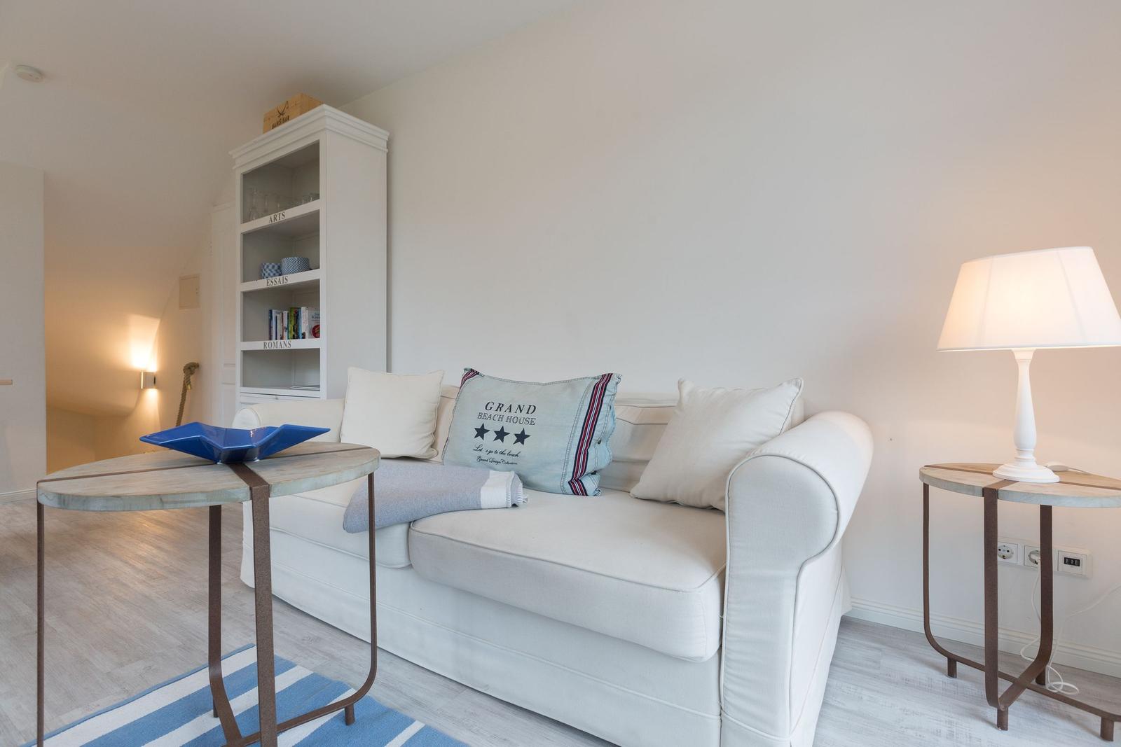 Weißes Sofa mit Kissen, Beistelltische und Bücherregal in einem hellen Raum.
