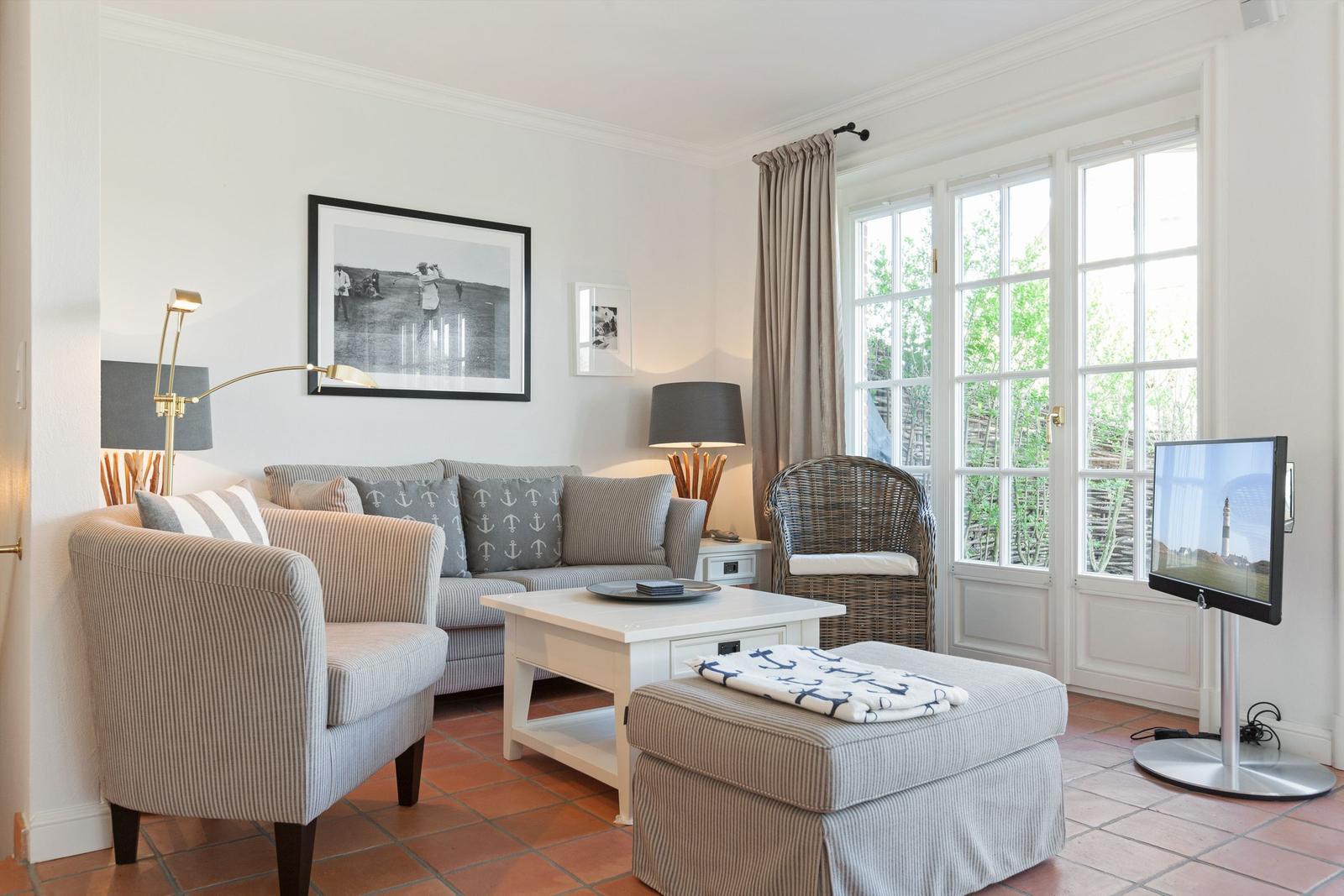 Wohnzimmer mit Sofa, Sessel, Couchtisch und Fernseher. Fenster mit Blick auf Garten.