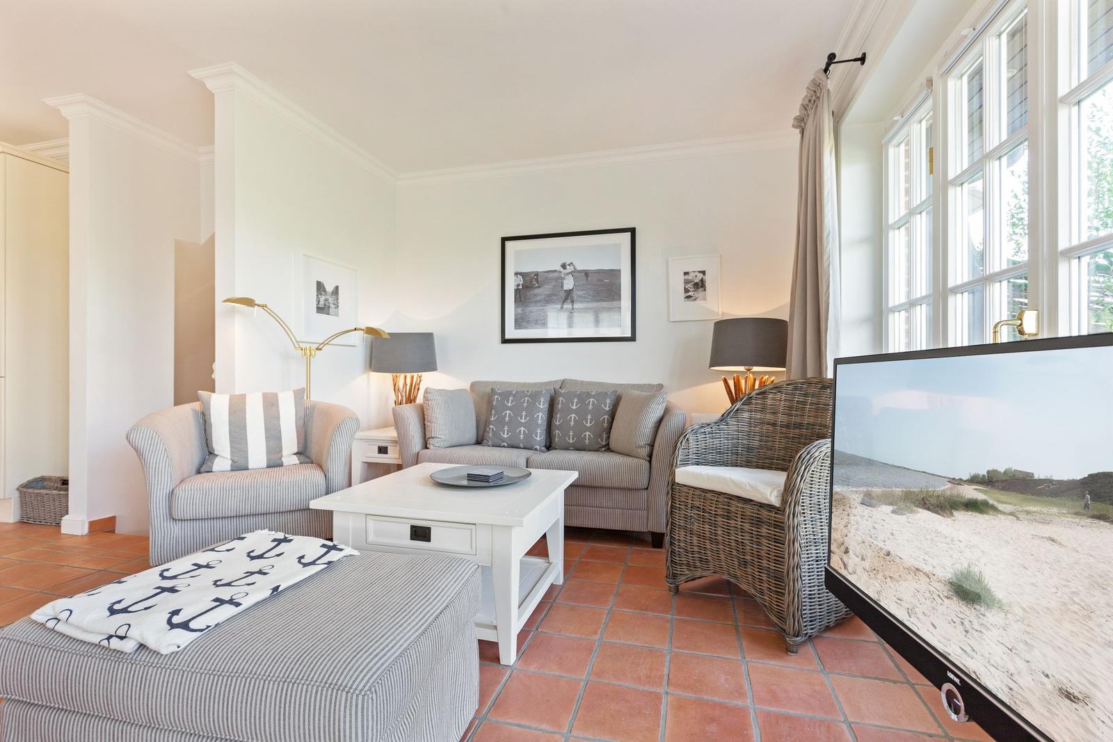Wohnzimmer mit Sofa, Sessel und Fernseher. Terrakottafußboden und große Fenster.