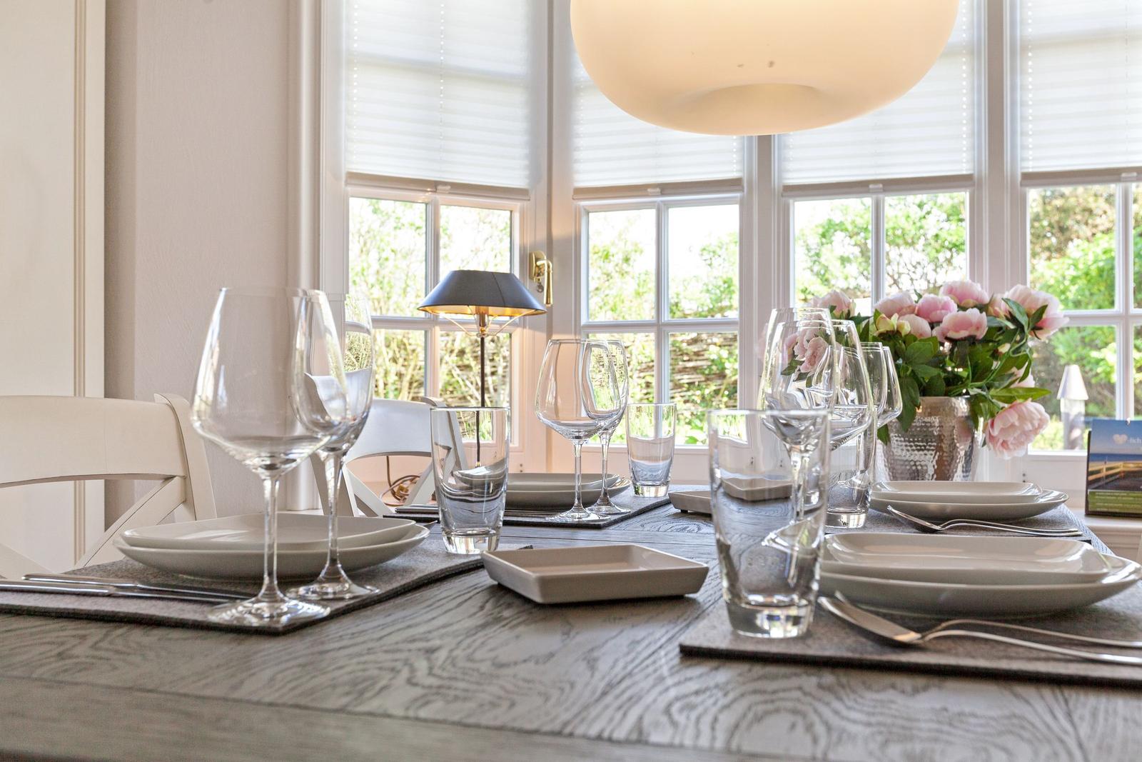 Gedeckter Esstisch mit Glas, Teller und Blumen vor Fenstern.