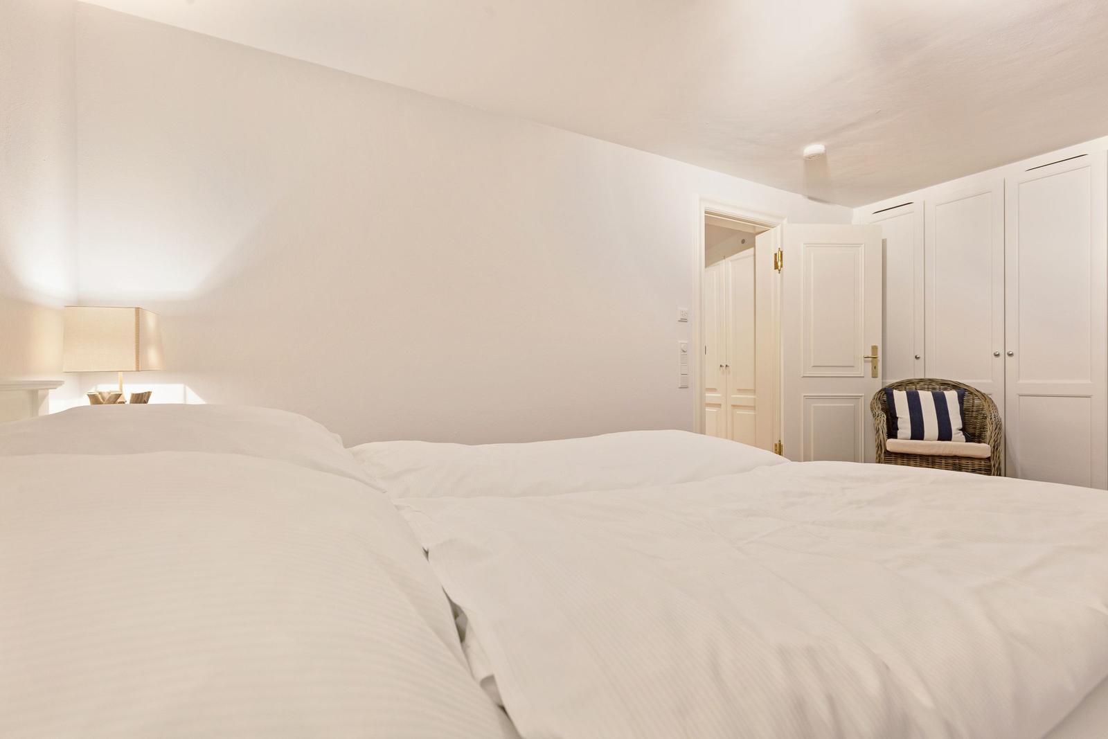 Schlafzimmer mit weißem Bett, Nachttischlampe und Stuhl mit Kissen.
